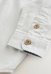 Lysfarvet bomuldsskjortemanchet med to runde, brune knapper. Pæn syning og en denimdetalje under manchetten.