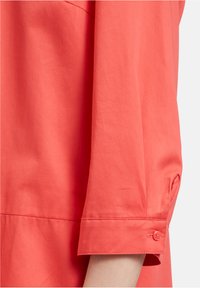 Koralrotet Hemd mit geknöpftem Manschette, das eine glatte Textur und einen subtilen Glanz aufweist. Der Ärmel ist am Handgelenk leicht ausgestellt.