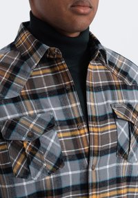 Geruite flanellen shirt in grijs, bruin en geel; voorzien van twee borstzakken, knoopsluitingen en een zachte textuur.
