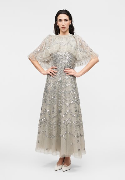 Γυναίκα με ασημί φόρεμα με παγιέτες, διαφανείς κοντές μανσέτες με βολάν και λευκά μυτερά τακούνια, στέκεται με τα χέρια στη μέση ενάντια σε λευκό φόντο.