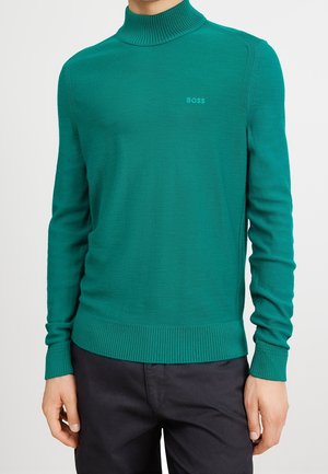 Muž nosící přiléhavý tyrkysový rolák se žebrovanými lemy rukávů a spodního okraje, doplněný tmavými kalhotami.