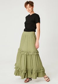Jupe maxi en tissu plissé vert clair avec des volants superposés au niveau de l'ourlet. Associée à un T-shirt noir ajusté et des sandales à lanières.