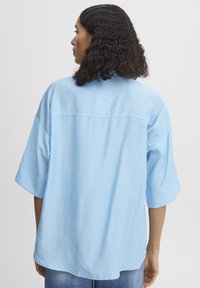 Camicia oversize azzurro chiaro con maniche corte, tessuto morbido, dettaglio della cucitura sulla schiena e orlo arrotondato, indossata con jeans scuri.