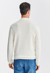 Kremowy sweter z dzianiny z prążkowanym kołnierzem i mankietami, o gładkiej strukturze i dopasowanym kroju, zestawiony z niebieskimi dżinsami.