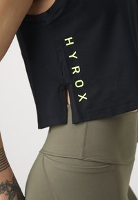 Svart kort tank top med sidöppning och neongul "HYROX"-tryck; matchas med figurnära olivgröna leggings med texturerade sömmar.