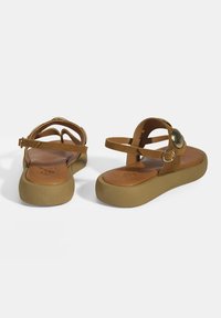 Braune Wildleder-Sandalen mit flacher Sohle und verstellbarem Knöchelriemen. Mit einem metallischen Akzent am oberen Riemen und einem konturierten Fußbett.