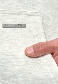 Tissu gris clair au toucher doux, avec une petite étiquette en argent portant la mention "bazix republiq" et une main pointant vers une poche.