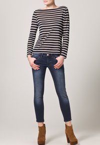 Žena oblečená v černo-bílém pruhovaném topu s dlouhým rukávem, tmavě modrých slim džínách a hnědých semišových vysokých kotníkových botách, stojící s rukama v kapsách.