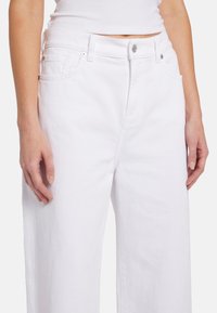 Pantaloni in denim bianchi a gamba larga con vita alta. Presentano tasche anteriori, chiusura con bottone e passanti per cintura. Tessuto liscio, dettagli minimi.