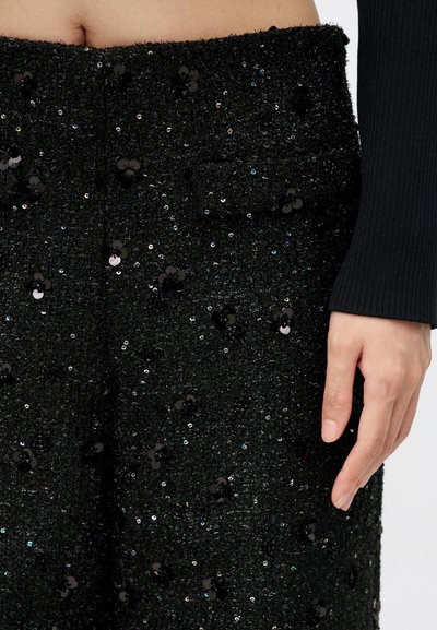 Pantalon taille haute texturé noir avec des motifs floraux en sequins noirs, détails de poche, et une main reposant sur le côté portant une manche à côtes noire.