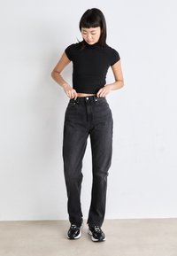 Svart ribbet crop top kombinert med høyt liv, mørk denim jeans. Modell iført svart-hvite sneakers, mot en ensfarget bakgrunn.