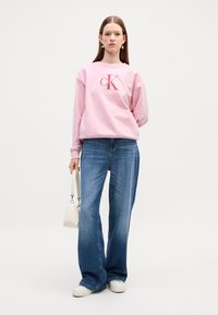 Felpa di cotone rosa chiaro con un grande logo rosso "CK", abbinata a jeans larghi blu e sneakers bianche. Il modello tiene una piccola borsa bianca.