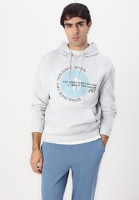 Sudadera gris claro con un bolsillo canguro, cordones y un gráfico circular en texto azul y negro. Combinada con pantalones azules.