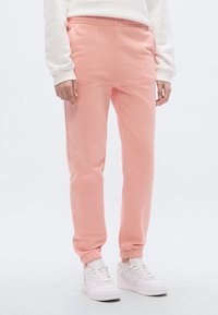 Calças de treino cor-de-rosa com cintura elástica e punhos canelados, feitas de tecido macio. Usadas com ténis brancos e uma camisola de cor clara.