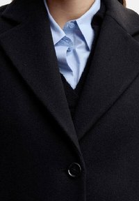 Close-up van een persoon die een zwarte knoopjas draagt over een lichtblauw shirt met kraag en een zwarte trui.