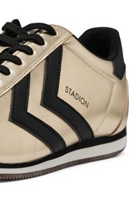 Närbild av guld- och svarta sneakers med svarta snören, chevronränder och texten "STADION" på sidan ovanför en vit och brun sula.