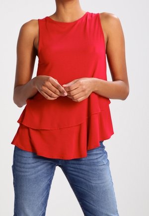 Femme portant un haut rouge sans manches superposé avec un jean bleu, debout, les mains doucement jointes devant un fond uni.