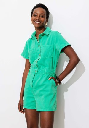 Femme souriante aux cheveux courts et bouclés porte une combinaison verte à manches courtes boutonnée avec de grandes poches, debout une main dans la poche.