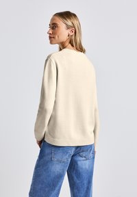 Beige gebreide trui met een ontspannen pasvorm, ribbelboorden en -zoom, gecombineerd met lichtblauwe jeans. De textuur lijkt glad en zacht.