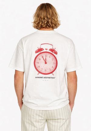 Persona con cabello rubio ondulado que lleva una camiseta blanca con un gran gráfico de un reloj despertador rojo y el texto "LEISURE AESTHETICS" en la parte posterior.