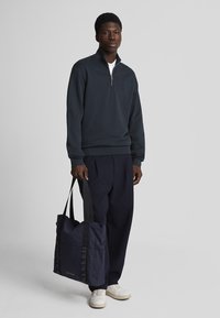 Donkerblauwe zip-up sweatshirt, katoenmix, geribbelde manchetten. Donkere broeken, lichte sneakers, met een grote donkerblauwe draagtas met gestructureerde hengsels.