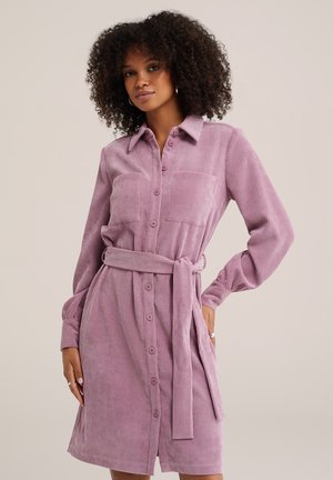 Femme aux cheveux bouclés portant une robe-chemise en velours côtelé mauve à manches longues avec boutons et ceinture nouée à la taille.