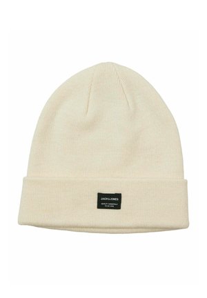 Cappello beanie crema realizzato in tessuto a maglia, con risvolto e un'etichetta nera con testo bianco sul fronte. Texture liscia.