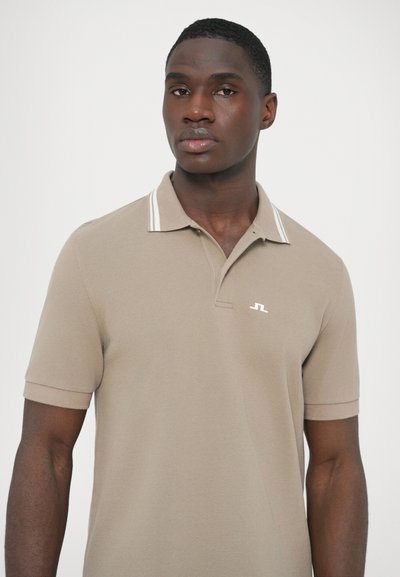 Μπεζ πόλο πουκάμισο από υφή υφάσματος, με γιακά με λευκές ρίγες. Ένα μικρό λογότυπο εμφανίζεται στην αριστερή πλευρά του στήθους.