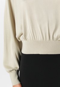 Beige Langarmoberteil mit gerafftem Saum und tailliertem Bündchen, kombiniert mit einem schlichten schwarzen Rock. Glatte Stoffqualität, schlichtes Design.