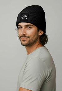 Black ribbed beanie with a rectangular patch displaying a white camera icon, worn by a person in a light gray shirt, close-up profile view.