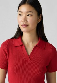 Röd ribbad polo-shirt med V-hals och korta ärmar. Tyget är texturerat, och kragen är enhetlig med en lätt vikning.