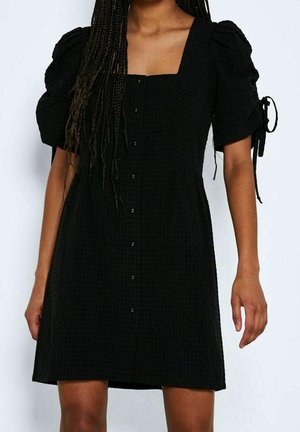 Kvinde iført en sort kortærmet kjole med knapper foran og puffede ærmer bundet med sløjfer.