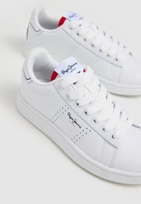 Scarpe sportive bianche realizzate in materiale sintetico con una finitura liscia. Le caratteristiche includono un design con lacci, accenti perforati e un logo sul lato.