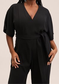 Combinaison noire avec un décolleté en V, des manches courtes et une taille ceinturée. Fabriquée en tissu lisse, elle possède des poches latérales et une coupe décontractée.