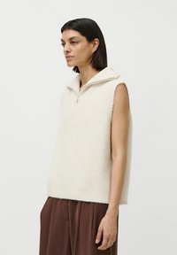 Femeie cu părul scurt și închis la culoare, purtând un vestă din tricot crem fără mâneci, cu guler cu fermoar și pantaloni maro lejeri, stând de profile.