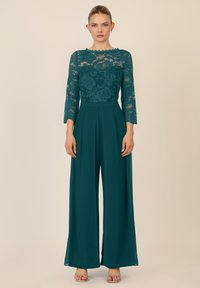 Tuta turchese con top in pizzo dotato di maniche a tre quarti, pantaloni a gamba larga plissettati e tacchi a spillo argentati. Tessuto liscio, design elegante.