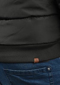 Fekete steppelt dzseki bordázott aljjal, barna bőr címkével. Kék farmernadrágon viselve, zseb részletekkel.