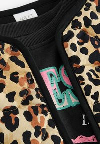 Chaleco acolchado con estampado de leopardo, con manchas beige, negras y marrones, combinado con una sudadera negra que muestra gráficos coloridos.