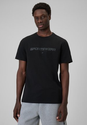 Mężczyzna w czarnej koszulce z napisem „SPORTSWEAR” i szarych dresach, stojący na jednolitym jasnoszarym tle.