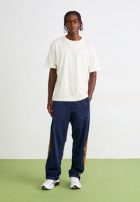 Mand med fletninger iført en off-white oversized t-shirt, marineblå træningsbukser med orange striber og hvide sneakers, som står på et grønt gulv.