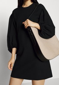 Robes noires à manches bouffantes en tissu lisse, avec un col rond. Le mannequin tient un sac à main structuré beige et noir.