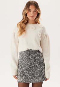 Vit, fluffig tröja med blommigt dekoration, i kombination med en kjol i silverfärgade paljetter. Outfiten har långa ärmar och en figurnära silhuett.