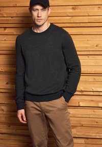 Dunkelgrauer Wollpullover mit rundem Ausschnitt und geripptem Saum, kombiniert mit hellbeigen Chinos. Der Hintergrund zeigt strukturierte Holzpaneele.