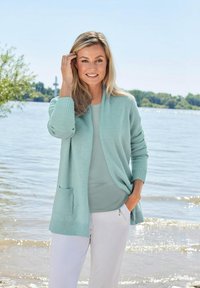 Hellblauer Cardigan aus weichem Material, mit offener Front, rippierten Bündchen und einer Vordertasche, kombiniert mit einem passenden Oberteil und weißen Hosen.
