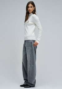 Vrouw die staat in een witte coltrui en losse grijze jeans, met zwarte puntige schoenen, tegen een effen lichtgrijze achtergrond.