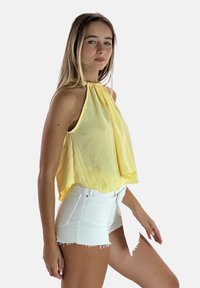 Top giallo a collare realizzato in tessuto leggero, con design plissettato. Abbinato a pantaloni shorts in denim bianco con effetto consumato, che mostrano orli sfrangiati.