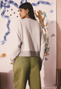 Hall hallipusa, millel on soonikkoesääris ja pikad varrukad, sobitatud rohelist värvi laiade pükstega, millel on tagataskud. Taustal on kapriissetest joonistustest.