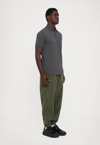 Man standing facing sideways wearing a grey short-sleeve polo shirt, green textured jogger pants, and black sneakers on a white background.