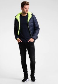 Sötétkék steppelt dzseki élénkzöld kapucnival, cipzáras lezárással. Tengerészkék ing és fekete farmer fölött viselve, fekete sportcipővel párosítva.