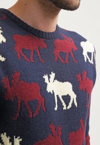 Maglione blu navy con motivi di alci in rosso e bianco. Tessuto a maglia strutturato con scollatura rotonda. Vista ravvicinata che mette in evidenza i dettagli del design.